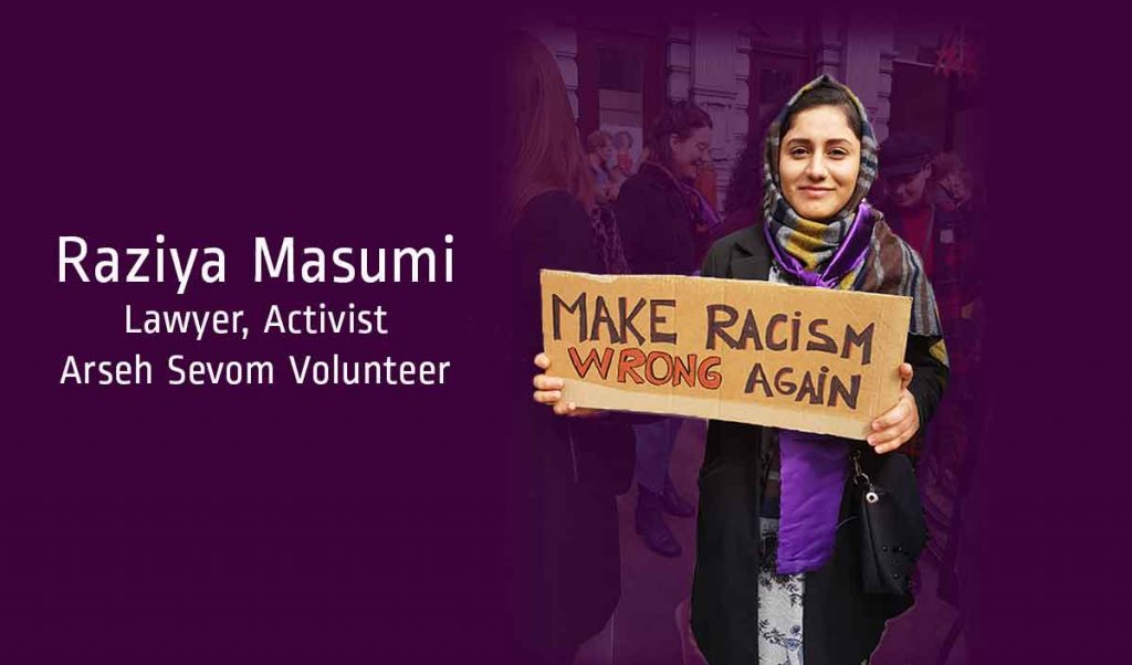 Young woman wearing a multi-colored scarf and holding a cardboard sign that reads "Make Racism Wrong Again"